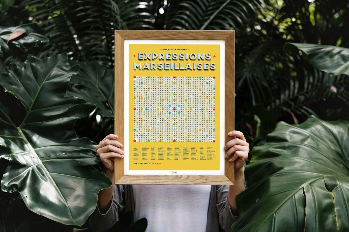 Affiche Expressions Marseillaises – mots cachés 100 mots – encadrée en bois naturel, tenue à bout de bras devant un fond végétal tropical