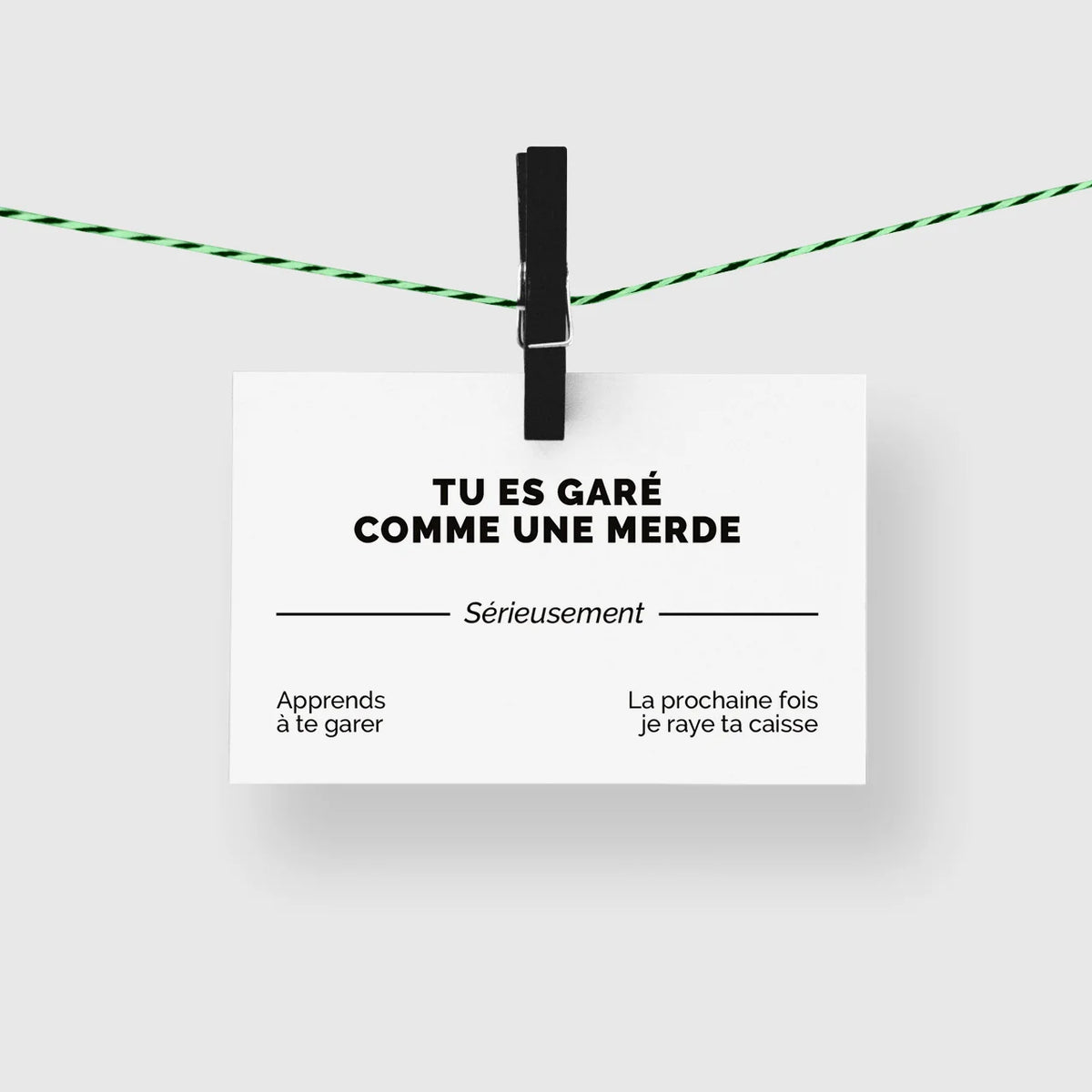 Carte de visite humour « Tu es garé comme une merde » suspendue à une corde à linge par une pince noire