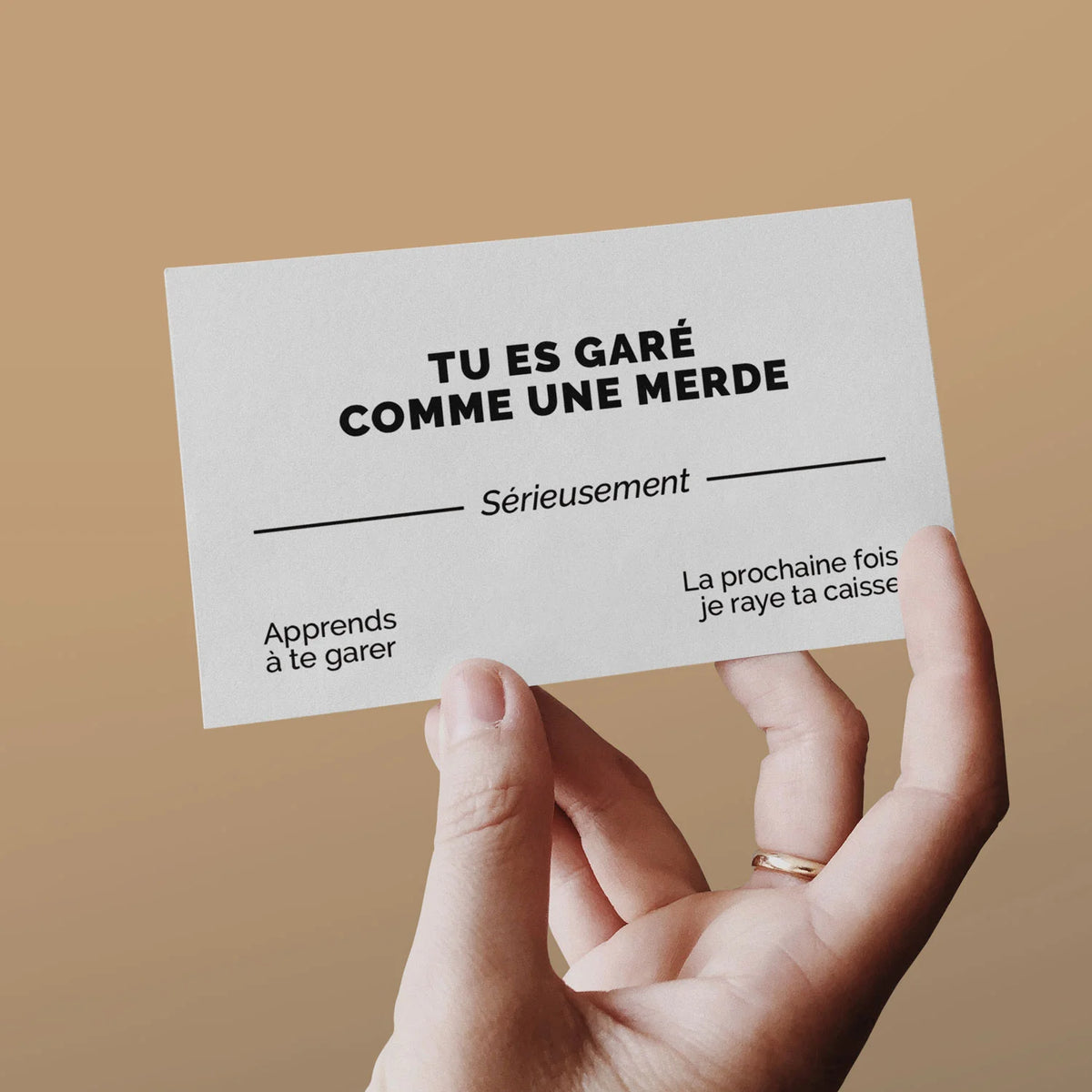 Main tenant une carte de visite « Tu es garé comme une merde » sur fond caramel