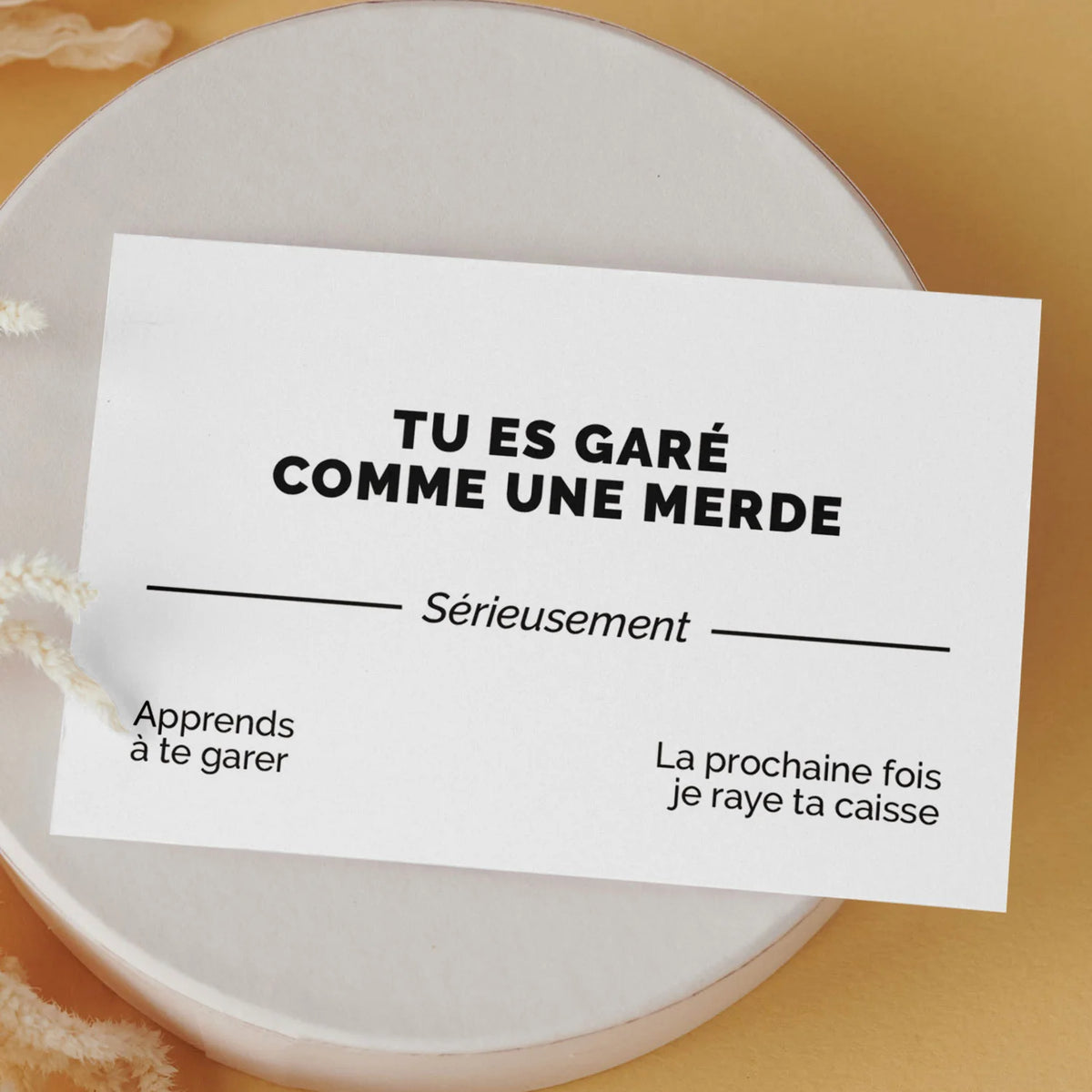Carte de visite drôle « Tu es garé comme une merde » posée sur une boîte ronde, fond orange